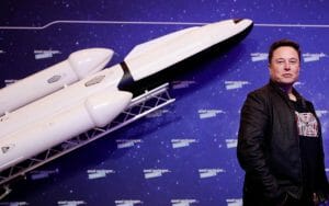 Tesla CEO Elon Musk is wearing a black jacket and standing next to an image of a spaceship