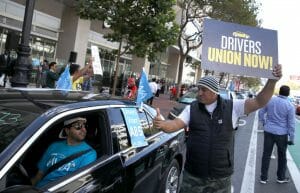 A man holding a sign with the slogan "drivers union now!" gives a thumbs up to a Uber driver.