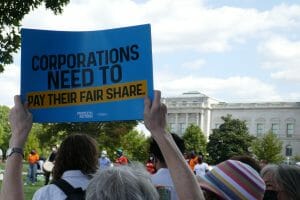 Person in a crowd is holding up a sign saying "Corporations Need to Pay Their Fair Share."