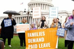 Five people are standing in front of the U.S. Capitol holding signs that read "Families and Paid Leave" and "Save Paid Leave."