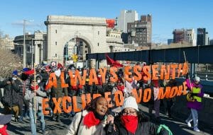 A march across a New York City Bridge with activists. An orange sign reads "Always Essential. Excluded no more."