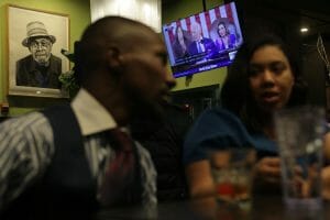Two people conversing in a bar with the State of the Union speech on a TV behind them.
