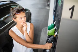 A woman wearing sunglasses and a white tank top looks concerned by gas prices while preparing to pump gas at the gas station.