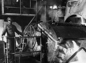 A Black man stands next to industrial equipment in a black and white image