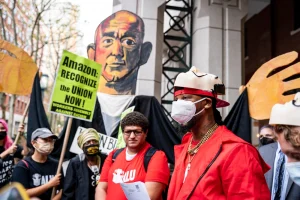 A man in a red shirt with a white mask speaks with a person holding a sign about labor rights behind him