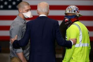 President Biden stands with his back to the camera, holding the arms of two workers.